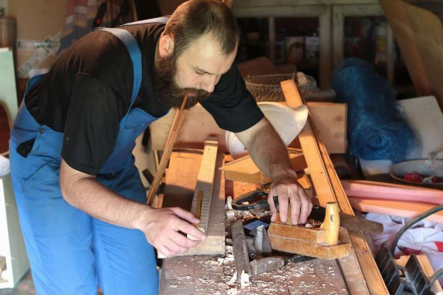 A bearded craftsman hand-planing a wood piece on a cluttered workbench, applying traditional frame joinery techniques in his workshop.