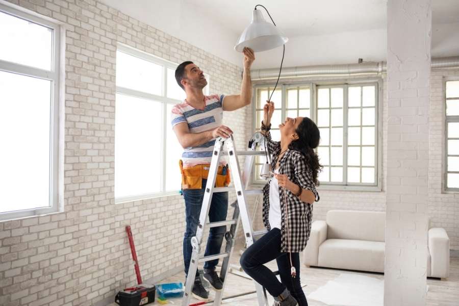 A couple working together on a ladder to install a new light fixture while renovating a fixer-upper.