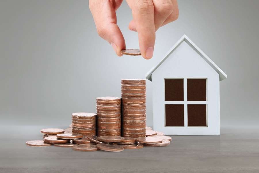 A hand placing a coin onto a stack next to a model house to negotiate the best price.