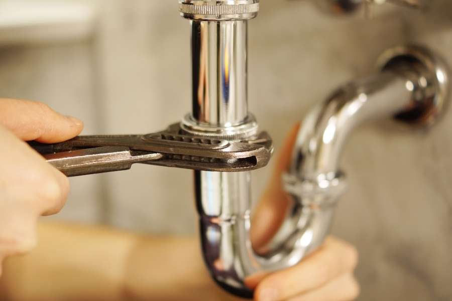 A close-up of hands using a wrench to tighten a chrome P-trap pipe assembly under a sink to prevent common plumbing mistakes.